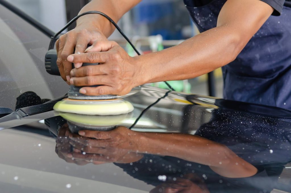 Detailed view of a workers hands polishing a cars hood using a buffer in an auto workshop.
