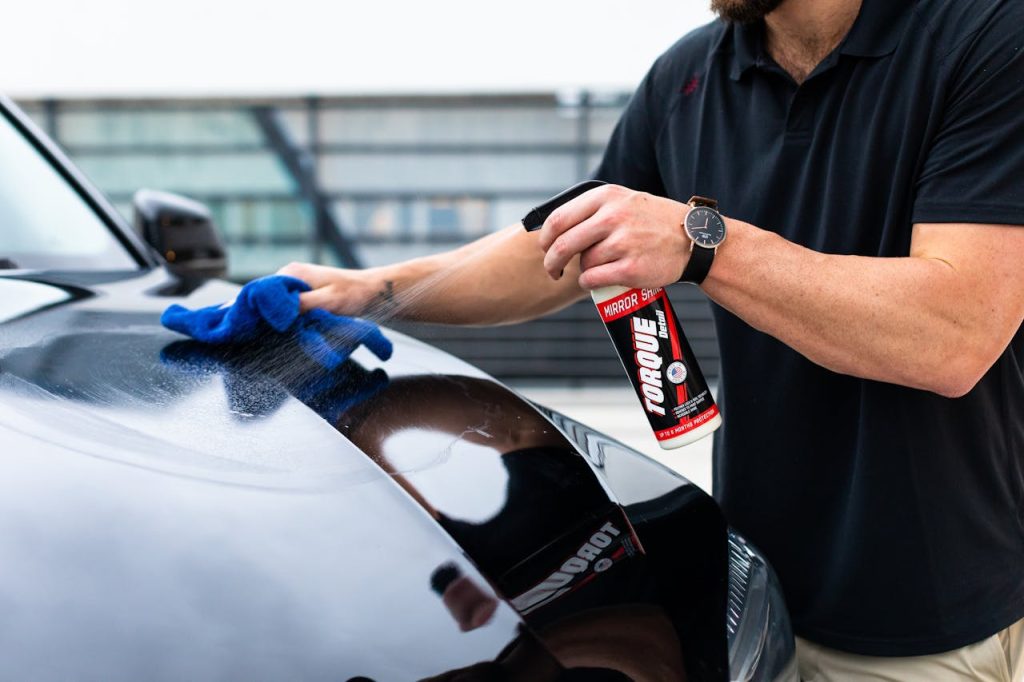 A man cleaning a black car hood with a spray and microfiber cloth.